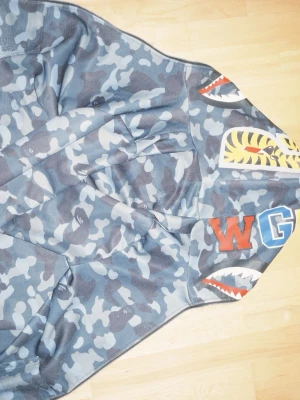 Blå camo hoodie från BAPE - Säljer en blå camo hoodie från BAPE med ikoniska haj- och tigertryck på huvan samt broderade bokstäver. Hoodien har dragkedja och cool streetwear-stil, perfekt för dig som vill sticka ut. Materialet är mjukt och bekvämt bomullstyg. Dragkedjan är sönder går att fixa hos skomakaren