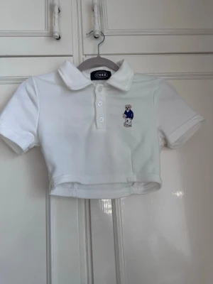 Vit croppad pikétopp från Cider - Säljer en vit croppad pikétopp från Cider med korta ärmar och krage. Toppen har knappar framtill och en söt björn brodyr på bröstet. Perfekt för en härlig sommardag! 💕