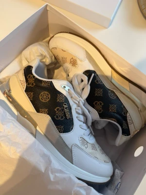 Guess sneakers - Snygga sneakers från Guess i vitt och beige med svarta paneler och guldiga logomönster. Skorna har vita snören, metallic-detaljer vid hälen och en klassisk låg siluett. Materialmix av läderimitation och textil för en trendig look.