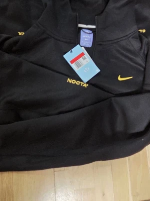 Svart NOCTA x Nike hoodie med gul logga - Svart hoodie från Nike NOCTA med gul Nike Swoosh och NOCTA-tryck på bröstet. Hoodien har huva och halv dragkedja framtill. Materialet är mjukt och sportigt, perfekt för en streetwear-look.