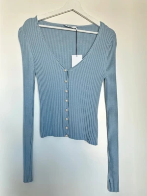 Ljusblå ribbad tröja med pärlknappar - Supersnygg ljusblå ribbad tröja med v-ringning och långa ärmar. Tröjan har pärlknappar framtill och är figurnära i modellen. Perfekt att styla med jeans eller kjol för en trendig look.