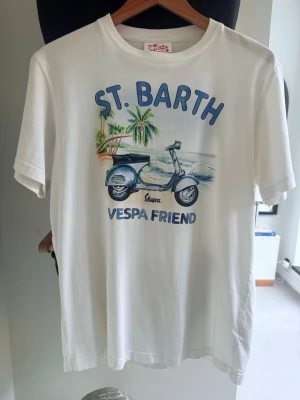Vit St. Barth Vespa t-shirt - Vit t-shirt från MC2 Saint Barth med tryck av en blå Vespa, palmer och texten 'ST. BARTH VESPA FRIEND' på framsidan. Klassisk passform och rund hals. Perfekt för en chill och avslappnad stil.