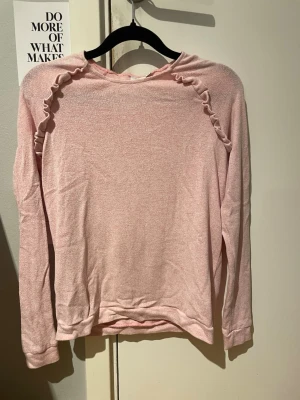Ljusrosa tröja med volangdetalj - Söt ljusrosa långärmad tröja med volangdetaljer längs axlarna. Tröjan har rund halsringning och är gjord i ett mjukt, tunt stickat material. Perfekt för en chill och feminin look. Skickar helst med PostNord😄