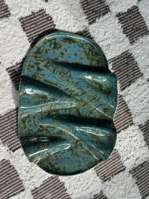 Charkbricka i keramik  - Säljer ett handgjort keramikfat med vågig form och glansig yta i blågröna och svarta nyanser. Materialet är keramik och färgerna skiftar snyggt i ljuset. Ca 40cm lång 25cm bredd 