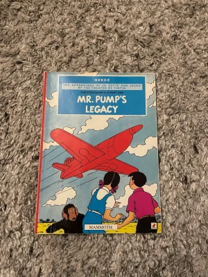 Mr. Pump's Legacy av Hergé - Engelskspråkigt seriealbum av Hergé med färgglada illustrationer på framsidan, där ett rött flygplan syns mot en blå himmel. Boken är en del av äventyren med Jo, Zette och Jocko och har ett mjukt omslag i papper.