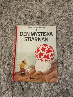 Den mystiska stjärnan Tintin-album - Säljer serieboken 'Den mystiska stjärnan' av Hergé ur serien Tintins äventyr. Omslaget är vitt med röd rygg och har en stor svamp med röda fläckar samt Tintin och Milou på framsidan. Boken är i klassiskt seriealbum-format och tryckt på papper.
