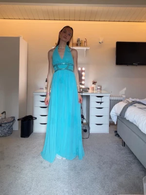 Turkos långklänning med halterneck - Säljer en turkos långklänning med halterneck och markerad midja med dekorativa pärlor. Klänningen har ett luftigt, svepande tyg och djup urringning framtill. Perfekt för dig som vill sticka ut med en färgstark och elegant look. Den är i nytt skick och aldrig använd 