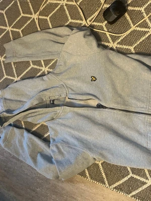  Lyle & Scott zip hoodie - Snygg grå hoodie från Lyle & Scott med dragkedja framtill och klassisk gul logga på bröstet. Tillverkad i mjuk bomullsblandning och har ribbade muddar vid ärmar och midja. Perfekt för en avslappnad stil.