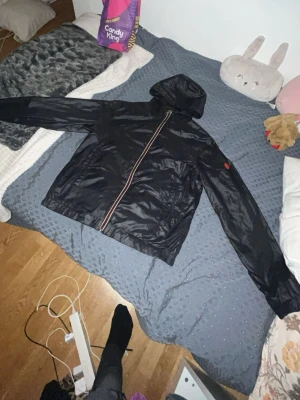 Svart vindjacka med huva - Svart vindjacka med huva och sneddragkedja framtill. Jackan har två sidofickor och en röd detalj på ena ärmen. Materialet är glansigt och lätt, perfekt för blåsiga dagar.