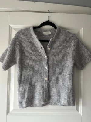 Grå ribbstickad kofta med kort ärm - Mysig grå ribbstickad kofta från Arket med korta ärmar och v-ringning. Koftan har knappar framtill i olika nyanser och är gjord i ett mjukt, luddigt material som känns lyxigt. Perfekt att slänga på sig över en t-shirt eller linne.