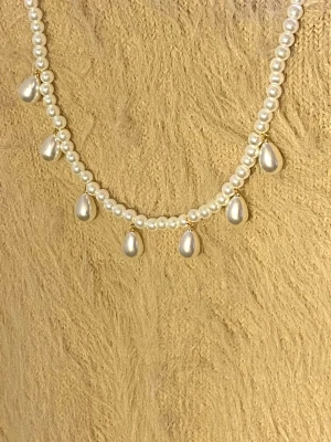 Pärlhalsband med droppformade hängen - Snyggt halsband med vita pärlor och sju droppformade pärlhängen. Halsbandet har en klassisk och elegant design med små runda pärlor och guldfärgade detaljer vid hänget. Perfekt för att lyfta din outfit med en stilren touch.