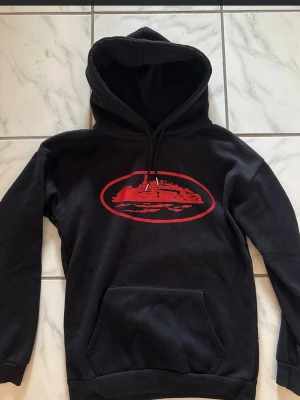 Svart hoodie med röd grafiskt tryck - Säljer en svart hoodie med huva och dragsnören. På bröstet finns ett stort rött grafiskt tryck av ett fartyg inuti en oval ram. Hoodien har en klassisk känguruficka framtill och ribbade muddar vid ärmslut och nederkant. Perfekt för dig som gillar streetwear och snygga prints.