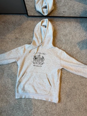 Grå hoodie från Bvaldi  - Snygg grå hoodie från Bvaldi med svart tryck på bröstet och stor ficka framtill. Klassisk huva med dragsko och mjukt, borstat innerfoder för extra komfort. Perfekt för en avslappnad och trendig stil.