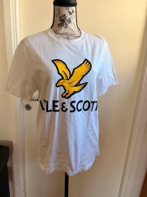 Vit Lyle & Scott t-shirt med tryck - Vit t-shirt från Lyle & Scott med stort gult fågeltryck och svart logga på bröstet. Klassisk rund hals och korta ärmar. Perfekt för en avslappnad och stilren look.