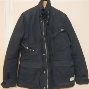 Hampton Republic jacka herr svart XL - Hampton Republic jacka för herr i storlek XL. Jackan är svart och har flera ytterfickor med dragkedja och knappar. Insidan har rutigt foder upptill och quiltat foder nedtill. Stängning med dragkedja och knappslå framtill samt hög krage med spänne. Passar som ytterjacka i vardagen eller till enklare utomhusaktiviteter.