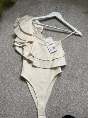 Krämvit body med volanger, Zara  - Supersnygg krämvit body från zara med en axel och dubbla volanglager över bysten och axeln. Tunn axelrem på ena sidan och hög skärning vid benen. Tillverkad i mjukt, lätt material som känns bekvämt mot huden. Perfekt för dig som vill ha något trendigt och unikt.