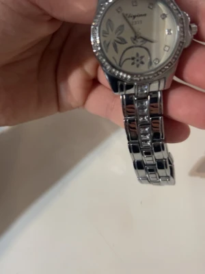 Silverfärgad klocka med blommönster - Stilren klocka från Elegance i silverfärg med metallarmband och glittrande stenar runt urtavlan. Urtavlan har ett blommigt mönster och markerade siffror med små stenar. Perfekt accessoar för dig som gillar detaljer.