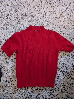 Röd ribbstickad topp med kort ärm stl xs - Snygg röd ribbstickad topp med korta ärmar och högre hals. Perfekt till jeans eller kjol för en trendig look. Materialet känns mjukt och stretchigt, vilket gör den bekväm att bära. Enkel men stilren design som passar till många outfits.