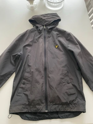 Grå vindjacka från Lyle & Scott - Snygg grå vindjacka från Lyle & Scott med huva och dragkedja framtill. Jackan har två fickor med  knapp, samt den klassiska gula loggan på bröstet. Jackan är använd och har ett litet minihål på framsidan men det är inget man lägger märke till.