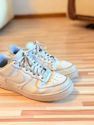 Vita Nike Air Force 1 sneakers - Säljer ett par klassiska vita Nike Air Force 1 sneakers med låg profil och snyggt lädermaterial. Skorna har perforeringar på tån för ventilation, ikonisk swoosh på sidan och platta vita skosnören. Perfekt för dig som gillar streetstyle.