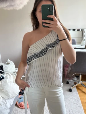 Randig oneshoulder topp med volang - Snygg vit randig oneshoulder topp med volangdetalj och svart mönster längs ned! Luftigt material och avslappnad passform.