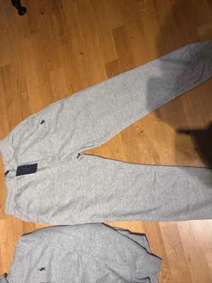 Grå mjukisbyxor från Polo Ralph Lauren - Snygga grå mjukisbyxor från Polo Ralph Lauren med klassisk logga broderad på vänster sida. Byxorna har resår i midjan och raka ben, tillverkade i mjukt bomullsmaterial för maximal komfort. Perfekta för chill dagar eller när du vill ha en avslappnad stil.