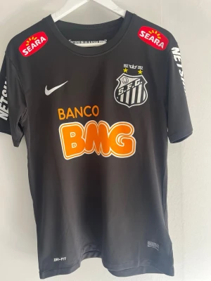 Svart Santos FC Neymar matchtröja - Svart fotbollströja för Santos FC med Neymar JR och nummer 11 på ryggen. Tröjan har flera sponsorloggor, klubbmärke på bröstet och är tillverkad i Dri-FIT-material som andas. Perfekt för fotbollsträning eller som supporterplagg.