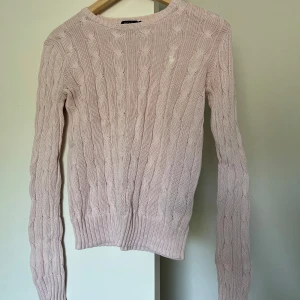 Ljusrosa kabelstickad tröja - Säljer en ljusrosa kabelstickad tröja med rund hals och långa ärmar. Tröjan har ribbstickade muddar vid ärmslut och nederkant. Perfekt för lager-på-lager och ger en klassisk look till din outfit.