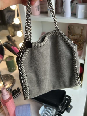 Grå axelväska med kedjedetaljer - Snygg grå handväska i mockaliknande material med kraftiga silverfärgade kedjor som handtag och längs kanterna. Väskan har en cool, modern look och passar perfekt till en trendig outfit. Kedjorna ger en edgy känsla och väskan är rymlig nog för det viktigaste.