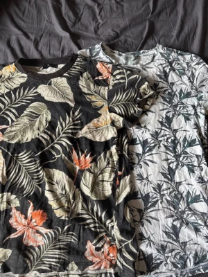 Svart t-shirt med blommigt mönster - Cool svart t-shirt med tropiskt blommigt mönster i beige, orange och grönt. T-shirten har korta ärmar och rund halsringning. Kontakta mig om du vill köpa en av dem 🥰