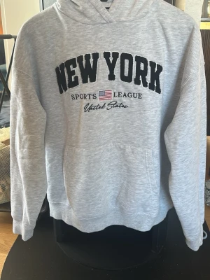 Grå hoodie  - Snygg grå hoodie från FB Sister i storlek XS med svart text 'NEW YORK' och broderad amerikansk flagga. Mjuk insida, huva och känguruficka framtill. Perfekt för en chill och sportig look.