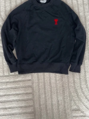 Svart sweatshirt från Ami Paris - Svart sweatshirt från Ami Paris med röd broderad logga i form av ett hjärta och bokstaven A på bröstet. Klassisk rund halsringning och ribbade muddar vid ärmslut och nederkant. Perfekt för en avslappnad och stilren look.