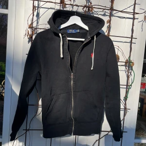 Svart hoodie från Polo Ralph Lauren - Säljer en svart hoodie från Polo Ralph Lauren med dragkedja, vit dragsko i huvan och den klassiska röda loggan broderad på bröstet. Hoodien har fickor framtill och är tillverkad i mjukt bomullsmaterial. Perfekt för en chill och stilren look.