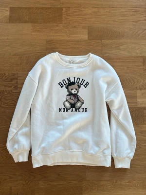 Vit sweatshirt med björntryck Springfield - Helt oanvänd vit sweatshirt från Springfield med ett gulligt björntryck och texten 'BONJOUR MON AMOUR' på bröstet. Tröjan har rund hals, ribbade muddar och är tillverkad i mjukt material som känns skönt mot huden. Perfekt för en avslappnad och trendig look.
