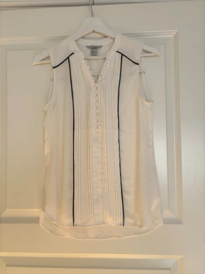Vit ärmlös blus med svarta detaljer - Snygg vit ärmlös blus med svarta ränder framtill och längs ryggen. Blusen har knappar och fina veckade detaljer. Storlek 36 och från H&M. Finns en liten fläck (se bild 3). 