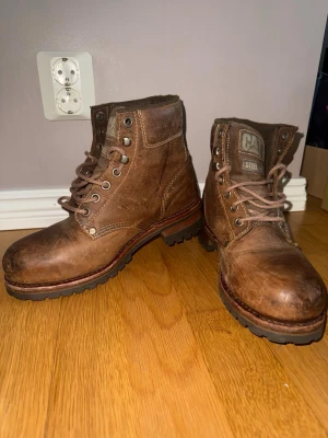 Bruna CAT kängor i läder - Tuffa bruna kängor från CAT med ståltå och grov sula. Tillverkade i slitstarkt läder med snörning och metallhål för extra hållbarhet. Perfekta för dig som vill ha en robust och snygg sko till vardagen. CAT-logga på plösen och undersidan.