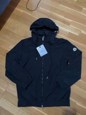 Svart Moncler vindjacka med huva - Snygg svart vindjacka från Moncler med huva och dragkedja framtill. Jackan har elastiska muddar, Moncler-logga på ärmen och diskreta rödvita detaljer vid huvan. Perfekt för blåsiga dagar och enkel att matcha med olika outfits. Komm med bud vid snabb affär