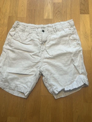 Beiga linne shorts från H&M - Snygga beige shorts i bomull med normal passform. De har bälteshällor, knapp och dragkedja framtill samt två sidofickor och två bakfickor med lock och knapp. Perfekta för varma dagar och enkel stil.