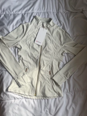 Vit jacka från Lululemon med dragkedja - Snygg vit sportjacka från Lululemon med figurnära passform, hel dragkedja framtill och två fickor med dragkedja. Jackan har långa ärmar och är tillverkad i ett stretchigt, mjukt material som passar perfekt till träning eller vardagsaktiviteter.
