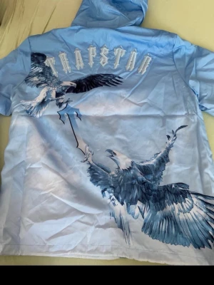 Blå Trapstar vindjacka med örntryck - Snygg blå vindjacka från Trapstar med stora örntryck på ryggen och logga i vitt. Jackan har huva och är tillverkad i ett lätt material som passar perfekt för blåsiga dagar. Jackan har en modern streetwear-look och cool design som sticker ut.