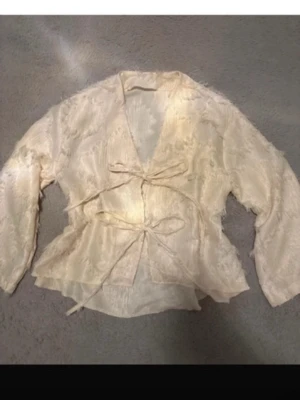 Krämvit omlottblus med knytband - Snygg krämvit omlottblus med två knytband framtill. Blusen har lång ärm och är gjord i ett glansigt, strukturerat tyg med fransiga detaljer. Perfekt för dig som gillar en unik och trendig look.