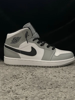 Air Jordan 1 Mid smoke grey  - Snygga Air Jordan 1 Mid sneakers i grått och vitt med svarta detaljer och svart swoosh. Skorna har klassisk Jordan-logga på sidan och är tillverkade i läder med perforerad tå för extra ventilation. Perfekta för dig som gillar streetwear och ikonisk design.