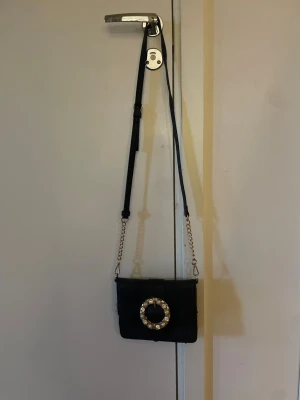 Svart axelväska med pärldetalj - Snygg svart axelväska i läderimitation med justerbar axelrem och guldfärgad kedja. Väskan har ett dekorativt spänne framtill med pärlor och gulddetaljer som ger en lyxig känsla. Perfekt för att lyfta din outfit.