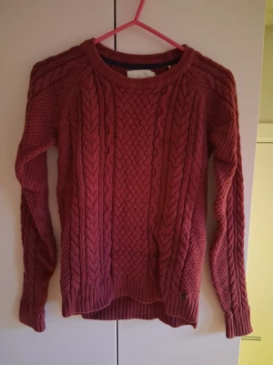 Vinröd kabelstickad tröja - Säljer en vinröd kabelstickad tröja med rund hals och långa ärmar. Tröjan har ett klassiskt flätmönster framtill och ribbade muddar vid ärmslut och nederkant. Perfekt för dig som gillar en mysig och stilren look.