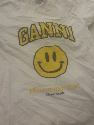 Vit Ganni t-shirt med smileytryck - Vit t-shirt från Ganni med stort gult smileytryck och texten 'Have a nice day! Please recycle' på framsidan. Klassisk passform och rund hals. Perfekt för dig som gillar statement-plagg med en lekfull touch.