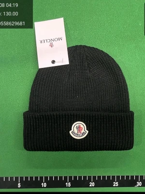 Svart Moncler mössa - Snygg svart ribbstickad mössa från Moncler med uppvikt kant och klassisk Moncler-logga framtill. Perfekt accessoar för att hålla värmen och ge din outfit en exklusiv touch. Tillverkad i mjukt och stretchigt material.