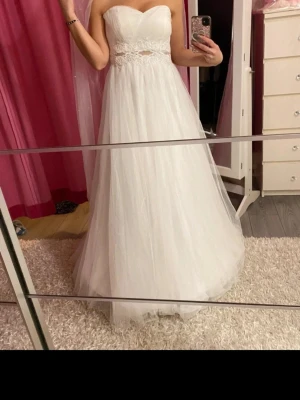 Vit långklänning med spetsdetaljer - Romantisk vit långklänning med axelbandslös design och vacker spetsdetalj i midjan. Klänningen har en lätt utsvängd kjol i flera lager tyg som ger en drömmig känsla. Perfekt för speciella tillfällen och student.Säljer en oanvänd vit klänning.  Kan användas på bröllop eller bal.  Dragkedja i ryggen Vadderad byst 2 lager under tyg och 2 lager tyll.  Fin detalj under bysten.  Extra axelband om man önskar med axelband.  Längd på hela klänningen ca 140 cm  Mått armhålan till armhålan 48 cm  2 underkj