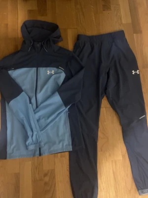Under Armour träningsset blå/grå - Snyggt träningsset från Under Armour med en blå och svart zip-jacka med huva och matchande svarta träningsbyxor. Jackan har logga på bröstet och ryggen, byxorna har logga på låret. Perfekt för löpning eller gym. Materialet är lätt och fun