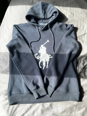 Mörkblå hoodie från Polo Ralph Lauren - Snygg mörkblå hoodie från Polo Ralph Lauren med stor vit polospelare och signatur på bröstet. Klassisk huva med dragsko och ribbade muddar vid ärmar och nederkant. Perfekt för en avslappnad och grisch stil. Matrialet är mjukt och bekvämt, perfekt för dagar då du vill klä dig grisch utan att tappa bekvämligheten. Säljer då jag har vuxit ur den.