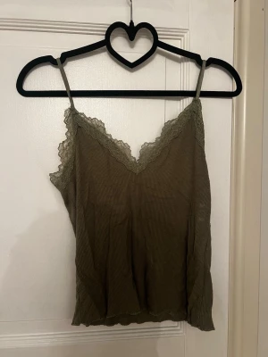 Olivgrönt ribbat linne med spets - Säljer ett olivgrönt linne med tunna axelband och v-ringning. Linnet har ribbad struktur och spetsdetaljer längs urringningen och upptill. Materialet känns mjukt och stretchigt. 